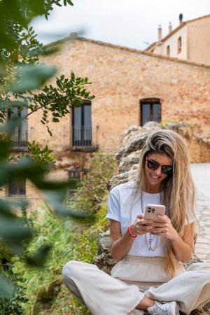 Téléchargez les photos : Femme blanche avec de longs cheveux blonds et des lunettes de soleil tapant avec son smartphone assis sur le mur près de la rivière - en image libre de droit