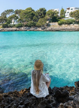 Téléchargez les photos : Vue arrière de la femme assise avec de longs cheveux blonds à Cala pi, Mallorca, îles Baléares. Eau turquoise arrondie par des rochers - en image libre de droit