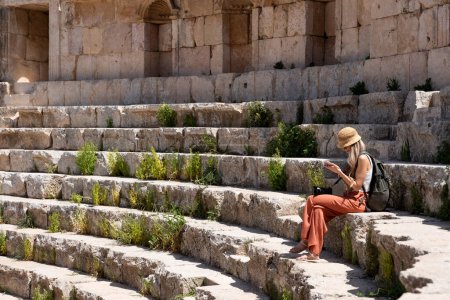 Téléchargez les photos : Voyage touriste femme blonde avec chapeau et sac à dos assis dans les escaliers en utilisant son smartphone dans le théâtre romain à Jerash, Ancienne ville romaine en Jordanie - en image libre de droit