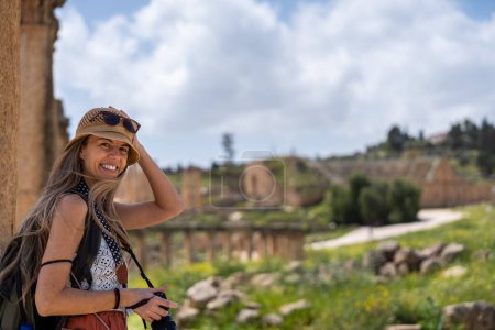 Téléchargez les photos : Femme blonde touristique avec photo, chapeau et cheveux longs sourit dans les ruines de l'ancien Jerash, Jordanie - en image libre de droit