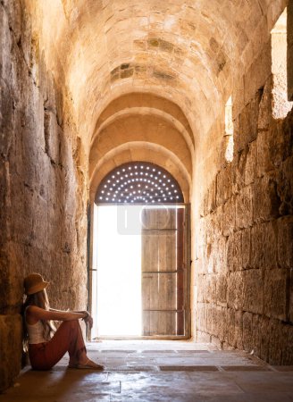 Téléchargez les photos : Vue arrière de la femme touristique blonde avec chapeau assis et soutenu sur le mur regardant la porte d'entrée. Ruines Jerash antiques, Jordanie - en image libre de droit
