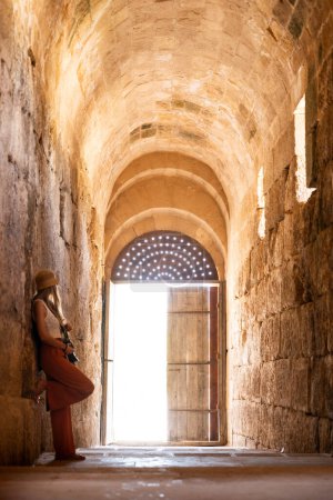 Téléchargez les photos : Vue arrière de femme touriste blonde avec chapeau soutenu sur le mur regardant la porte d'entrée avec photographie caméra Ruines Jerash antiques, Jordanie - en image libre de droit