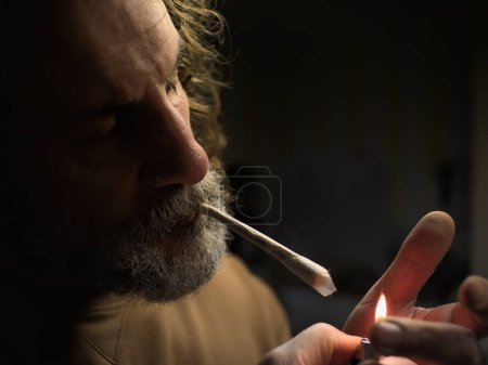 Foto de Hombre barbudo con el pelo largo está encendiendo un cigarrillo cónico grande o una articulación de la droga con un encendedor. - Imagen libre de derechos