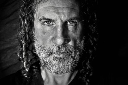 Photo for Contrasting monochrome frontal portrait of a Caucasian man in his forties, with long curly hair and a black and white beard. He is looking directly at the lens. Black background. - Royalty Free Image