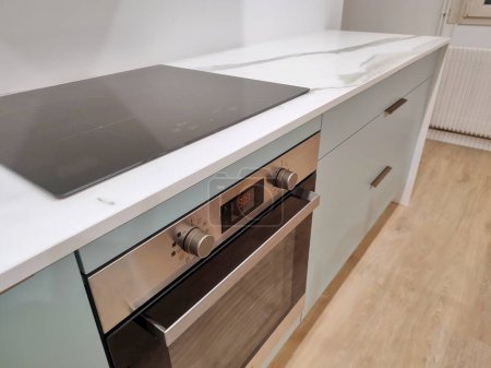 Marbled ceramic countertop, set on a blue-green kitchen cabinet with bronze handles. An induction hob and oven have been integrated.
