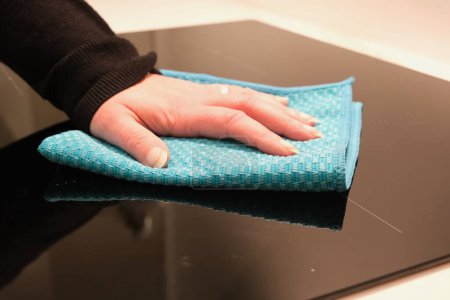 Woman's hand holding a blue microfiber cloth and cleaning an induction hob.