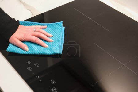 Woman's hand holding a blue microfiber cloth and cleaning an induction hob.