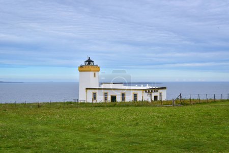 Photo for White lighthouse at Duncansby Head, green fields and blue ocean under a cloudy summer sky. Beacon in Scotland, United Kingdom, travel Europe - Royalty Free Image