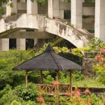A bamboo-framed gazebo with a black coconut fiber roof, nestled in a garden against the backdrop of a large highway bridge structure, offering a tranquil resting spot amid urban hustle.