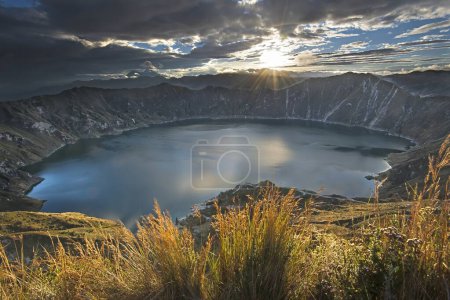 Téléchargez les photos : Dramatic sunrise shot of the volcanic caldera and Quilotoa lake and crater. The sun rises over the caldera and reflects on the calm dark water - en image libre de droit