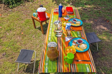 Téléchargez les photos : Tabourets de camping et table de pique-nique dans le camping forestier avec eau douce, jus, légumes, salade de fruits, haricots, omelette et pain grillé - en image libre de droit