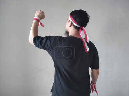 Téléchargez les photos : Vue arrière d'un Indonésien portant une chemise noire et un bandeau de drapeau indonésien levant les bras et serrant les poings - en image libre de droit