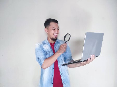 Photo for Mature Asian man looking at his laptop through a magnifying glass with happy expression - Royalty Free Image