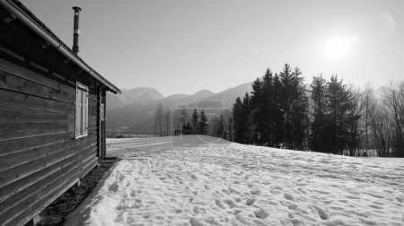 Foto de Cabaña de madera con persianas y chimenea estrecha en la nieve donde se pueden ver pasos y rayos de sol, bosques y montañas en el fondo en blanco y negro en Austria - Imagen libre de derechos