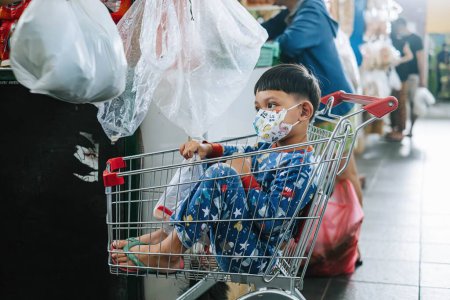 Photo for Hi-res photo of an Indonesian kid who wear mask is sitting on the shopping cart - Royalty Free Image