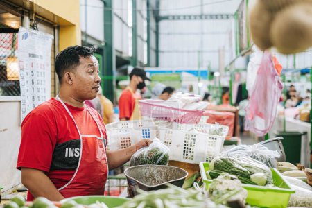 Foto de Foto de Hi-res de un vendedor indonesio que usa camiseta roja y delantal está vendiendo comestibles en el mercado tradicional con bokeh - Imagen libre de derechos