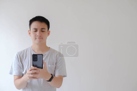 Foto de Un joven asiático que lleva una camiseta gris y un reloj inteligente sostiene y mira el teléfono inteligente con confusión - Imagen libre de derechos