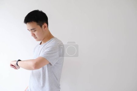 Foto de Un joven asiático vistiendo una camiseta gris está comprobando el reloj en un reloj inteligente en su muñeca izquierda en un fondo blanco aislado - Imagen libre de derechos