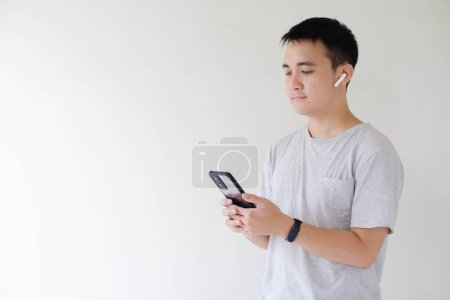 Foto de Un joven asiático que usa una camiseta gris, usando un par de auriculares inalámbricos y un reloj inteligente en su muñeca izquierda sostiene y mira un teléfono inteligente mientras escucha música en sus auriculares inalámbricos. Fondo blanco aislado. - Imagen libre de derechos