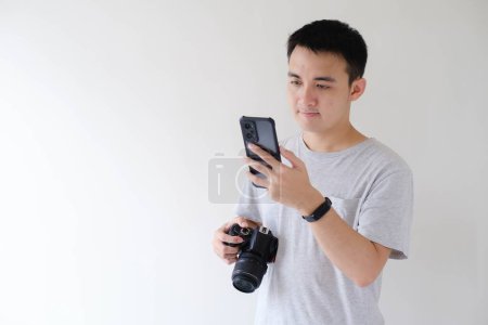 Foto de Un joven asiático con una camiseta gris está mirando un teléfono inteligente en su mano izquierda y sosteniendo una cámara réflex digital en su mano derecha. Fondo blanco aislado. - Imagen libre de derechos