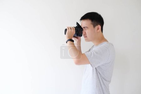 Foto de Un joven asiático con una camiseta gris y un reloj inteligente en la muñeca izquierda está fotografiando y dirigiendo una cámara réflex digital hacia el lado izquierdo con el flash abierto. Fondo blanco aislado. - Imagen libre de derechos