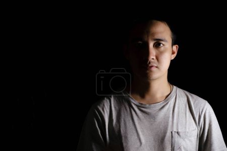 Foto de Una foto de bajo perfil de un joven asiático con una camiseta gris y un collar de metal plateado está mirando a la cámara con una cara parcial en la sombra. Fondo negro aislado. Dramático.. - Imagen libre de derechos
