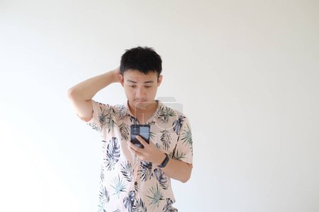 Foto de Un joven asiático que lleva una camisa rosa con patrones tropicales se confunde mientras sostiene un teléfono inteligente. Mano de la cabeza. Fondo blanco aislado. - Imagen libre de derechos