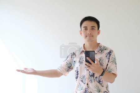 Foto de Un joven asiático vistiendo una camisa rosa con patrones tropicales y un reloj inteligente en su muñeca izquierda. Su mano izquierda sostiene un smartphone, su mano derecha es la palma de la mano abierta. Fondo blanco aislado. - Imagen libre de derechos