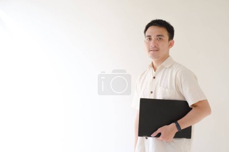Photo for A young Asian man wearing a beige shirt and a smartwatch on his left wrist is carrying a black laptop while looking at the camera. Isolated white background. Suitable for advertisement. - Royalty Free Image