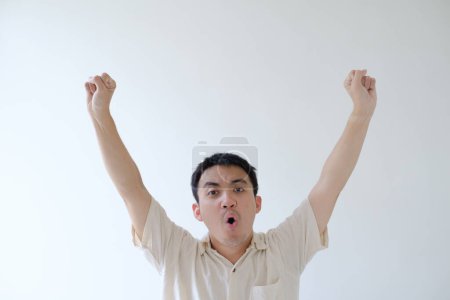 Foto de Un joven asiático con una camisa beige está levantando ambas manos con una expresión facial excitada. Fondo blanco aislado. - Imagen libre de derechos