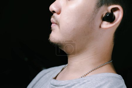 Foto de Primer plano de la cara parcial de un joven asiático que llevaba una camiseta gris y escuchaba audífonos inalámbricos negros redondeados. Enfocado en los auriculares. Fondo negro aislado. - Imagen libre de derechos