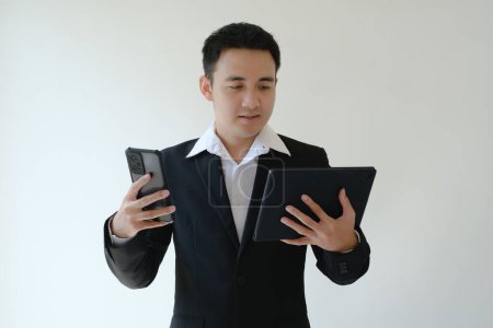 Foto de Joven empresario asiático sosteniendo un teléfono inteligente y un dispositivo de tableta y mirándolo. Fondo blanco aislado. - Imagen libre de derechos