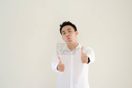 Foto de Joven asiático vestido con una camisa blanca y un reloj le da dos pulgares a la cámara con una expresión facial divertida. Fondo blanco aislado. - Imagen libre de derechos