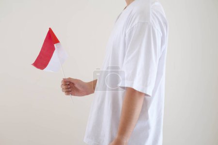 Foto de Un hombre que lleva una camiseta blanca sostiene la bandera de Bendera Indonesia o Indonesia mientras mira hacia el lado izquierdo sobre un fondo blanco aislado. - Imagen libre de derechos