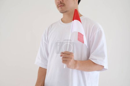 Foto de Un hombre vistiendo una camiseta blanca sostiene la bandera de Bendera Indonesia o Indonesia sobre un fondo blanco aislado. - Imagen libre de derechos