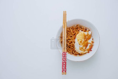 Téléchargez les photos : Plan plat ou vue de dessus d'une portion de nouilles instantanées indonésiennes sautées avec oeuf sur un bol blanc avec une paire de baguettes en bois sur une table blanche - en image libre de droit