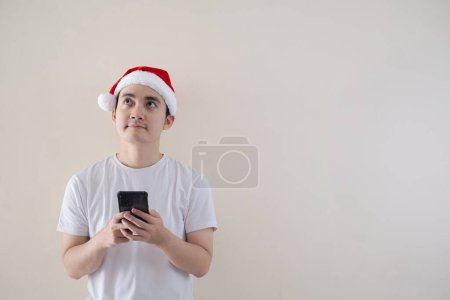 Foto de Joven asiático Santa hombre se confunde mientras sostiene un teléfono inteligente y mirando hacia arriba. Fondo beige aislado. Concepto de Navidad. - Imagen libre de derechos