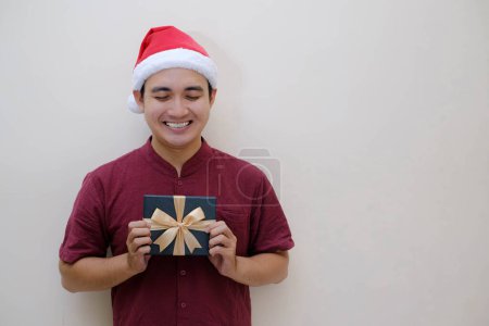Foto de El joven asiático Santa está sosteniendo una caja de Navidad verde esmeralda con expresión de sonrisa. Fondo beige aislado. - Imagen libre de derechos