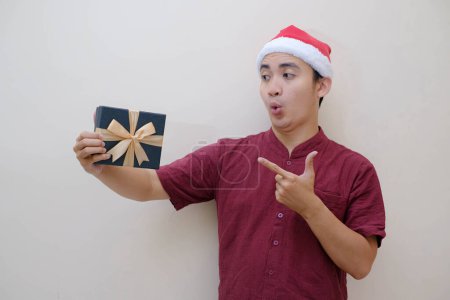 Foto de Joven asiático Santa hombre está sosteniendo y mostrar una caja de Navidad verde esmeralda a la cámara con expresión de la cara conmocionada. Fondo beige aislado. - Imagen libre de derechos