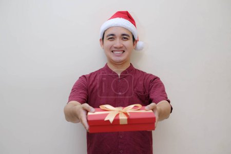 Foto de Joven asiático Santa hombre está dando una caja de regalo roja a la cámara con la sonrisa expresión de la cara. Fondo beige aislado. Concepto de Navidad. - Imagen libre de derechos