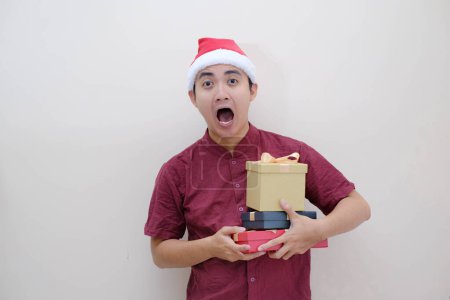 Foto de Joven asiático Santa hombre está sosteniendo cajas de Navidad con la expresión de la cara impactada y mirando a la cámara. Fondo beige aislado. Concepto de Navidad. - Imagen libre de derechos