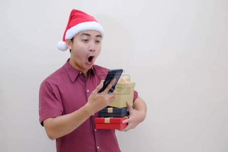 Foto de Joven asiático Santa hombre está sosteniendo cajas de Navidad con expresión de la cara impactada mientras mira el teléfono inteligente. Fondo beige aislado. Concepto de Navidad. - Imagen libre de derechos