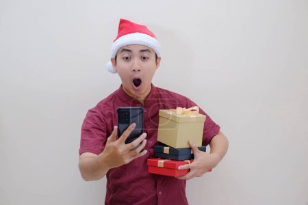Photo for Young Asian Santa man is holding Christmas boxes with shocked face expression while looking at the smartphone. Isolated beige background. Christmas Concept. - Royalty Free Image