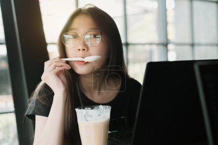 Foto de Joven mujer asiática está cucharando ingredientes de un vaso de café mientras mira a la cámara delante de su computadora portátil por la tarde - Imagen libre de derechos