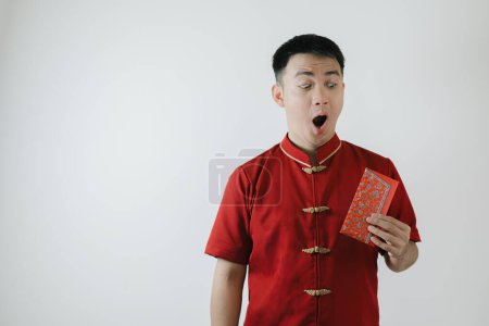 Téléchargez les photos : Visage surpris de l'homme asiatique portant un tissu traditionnel chinois appelé Cheongsam tenant un angpao sur fond blanc - en image libre de droit