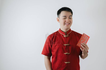 Téléchargez les photos : Visage souriant de jeune homme asiatique portant un tissu traditionnel chinois appelé Cheongsam tenant un angpao sur fond blanc - en image libre de droit