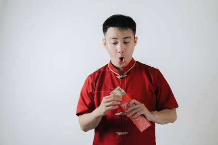 Téléchargez les photos : Visage choqué de l'homme asiatique portant un tissu traditionnel chinois appelé Cheongsam tout en tirant des sommes d'argent d'un angpao sur fond blanc - en image libre de droit