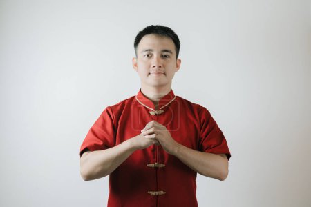 Téléchargez les photos : Geste de félicitations de l'homme asiatique portant un tissu traditionnel chinois appelé Cheongsam sur le Nouvel An lunaire et en regardant la caméra sur fond blanc - en image libre de droit