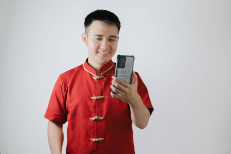 Téléchargez les photos : Visage souriant de l'homme asiatique portant un tissu traditionnel chinois ou Cheongsam tout en regardant son smartphone sur fond blanc - en image libre de droit
