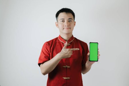 Téléchargez les photos : Asiatique homme portant tissu traditionnel chinois ou Cheongsam pointant vers son smartphone écran vert et regardant la caméra sur fond blanc - en image libre de droit